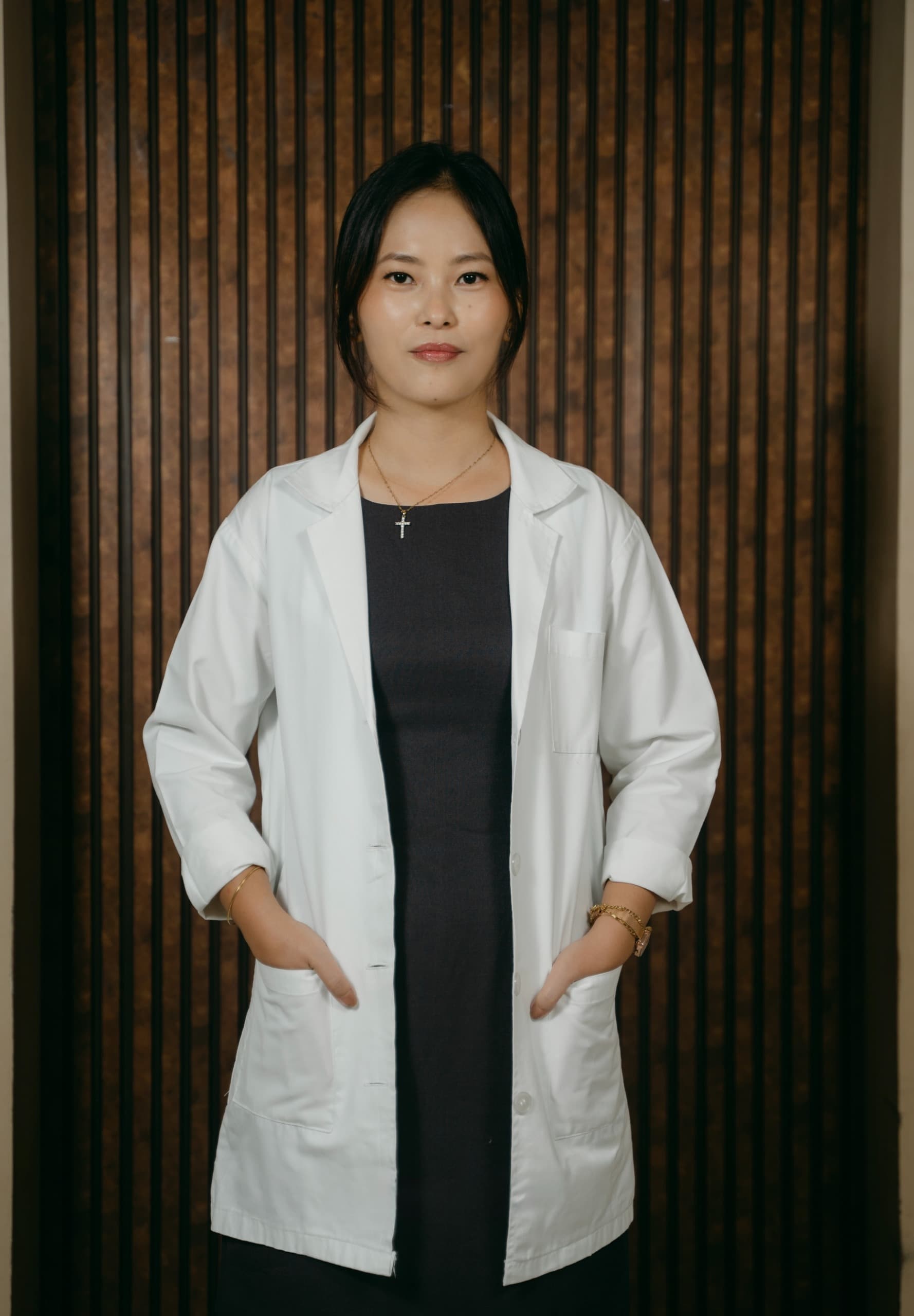 Professional woman in a white lab coat standing with hands in pockets against wooden slats.